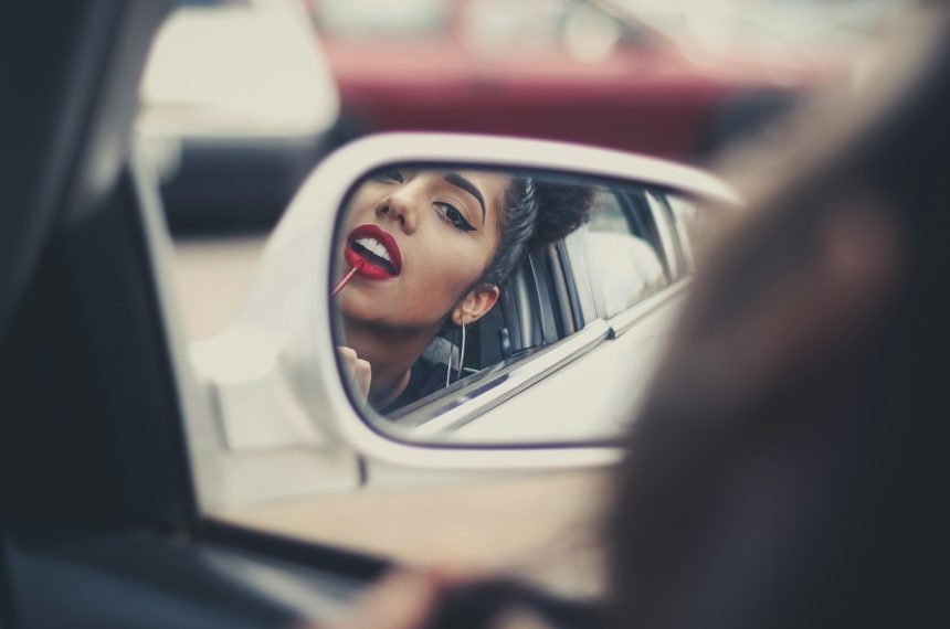 woman putting liquid lipstick on her lips while looking at vehicle's mirror during daytime